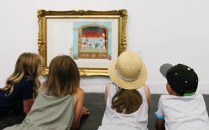 Czworo dzieci leży na podłodze i ogląda ilustrację I. Chmielewskiej oprawioną w złotą ramę.