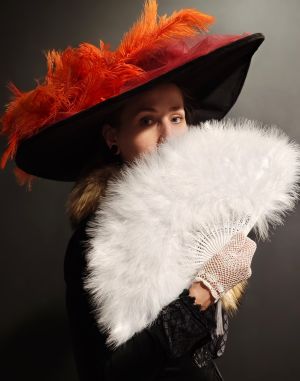 Kobieta w kapeluszu z piórami, trzymająca w dłoniach wachlarz, na rękach ma koronkowe rękawiczki.
