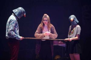 Troje młodych aktorów na scenie. Dwie osoby mają maski ptaków na głowach.