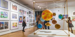 Na ścianach kolorowe obrazy, na pierwszym planie instalacja układu planetarnego, w tle postaci dzieci i dorosłych