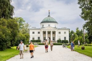 Biały zabytkowy budynek z zieloną kopułą wśród drzew.