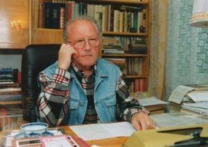 Mężczyzna w okularach siedzi przy biurku, na którym leży stos różnych notatek, dokumentów, książek.