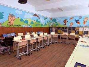 Kolorowa sala z ławkami szkolnymi ustawionymi w kształt podkowy, w tyle szafki z akcesoriami.