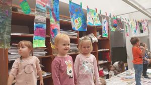 Uśmiechnięte małe dzieci stoją przy kolorowych pracach plastycznych.