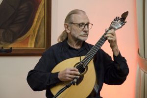 Mężczyzna o długich jasnych włosach i w okularach gra na gitarze portugalskiej.