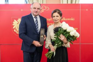 Marszałek województwa ze statuetką stoi obok liderki edukacji trzymającej bukiet białych róż.