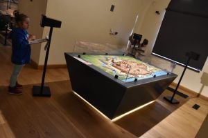 Małe dzieci oglądają makietę miasta w muzeum; jedno z nich trzyma kartkę i ołówek, robiąc notatki.