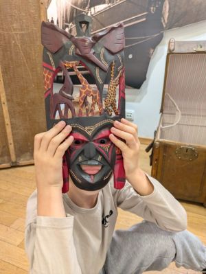 Dziecko w jasnych ubraniach trzyma kolorową, afrykańską maskę z motywami zwierząt, zasłaniając nią twarz.