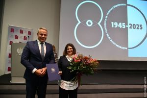 Prezydent miasta w garniturze i dyrektorka szkoły z bukietem kwiatów. W tle duże logo jubileuszu.