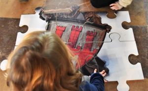 Dziecko układa duże puzzle na podłodze, przedstawiające herb z czerwonymi wieżami.