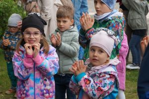 Grupa dzieci na dworze, ubrane w kolorowe kurtki i czapki, klaszcze w dłonie podczas wspólnej zabawy.