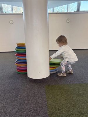 Małe dziecko układa kolorowe, okrągłe poduszki obok białej kolumny w jasnym, nowoczesnym wnętrzu.