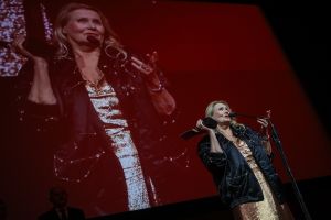 Piękna kobieta w stroju wieczorowym przemawia na scenie, w podniesionej ręce trzyma statuetkę.