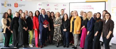 Grupa elegancko ubranych kobiet i mężczyzn pozuje do zdjęcia na tle bannera akcji.