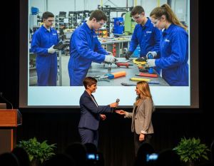 W auli kobieta przekazuje mikrofon drugiej mówczyni. W tle ekran ze zdjęciem uczniów w warsztacie.