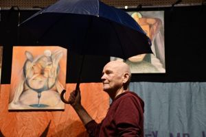 Aktor na scenie trzyma otwarty parasol. W tle obrazy abstrakcyjne.