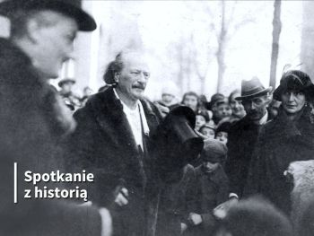 Ignacy Paderewski trzyma cylinder w dłoni. Wokół niego skupieni są dorośli w kapeluszach i dzieci.