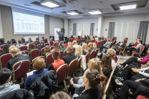 Uczestnicy konferencji w sali z dużym ekranem, na którym wyświetlane są dane statystyczne.