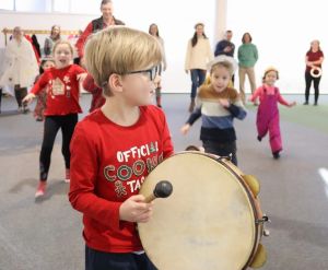 Dzieci bawią się w sali; chłopiec z przodu gra na bębnie, a reszta z radością biegnie za nim.