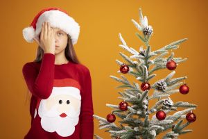 Dziewczynka w świątecznym stroju stoi obok choinki, z poważną miną zakrywa ręką połowę twarzy.