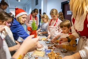 Dzieci w fartuszkach kuchennych dekorują świąteczne pierniczki stojąc wokół stołu z choineczką.