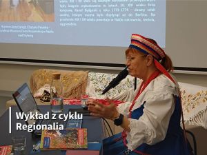 Kobieta w stroju regionalnym przy laptopie podczas wykładu. Na dużym ekranie wyświetlana jest prezentacja.