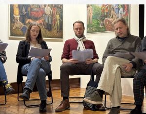 Grupa osób siedzi na krzesłach i czyta teksty podczas spotkania, w tle obrazy w galerii.