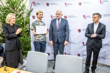 Kobieta i trzech mężczyzn stojących w urzędzie, jeden z mężczyzn uśmiecha się i pokazuje dokument.