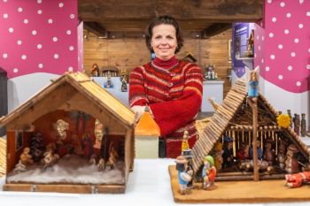 Uśmiechnięta kobieta stoi między drewnianymi szopkami bożonarodzeniowymi, w tle inne szopki.