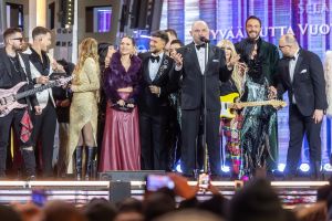 Na scenie elegancko ubrani  kobiety i mężczyźni z instrumentami muzycznymi, przy mikrofonie przemawia mężczyzna.