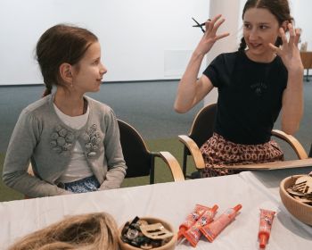 Uśmiechnięte dziewczynka i młoda kobieta z nożyczkami w uniesionej ręce siedzą przy stole z przyborami plastycznymi.