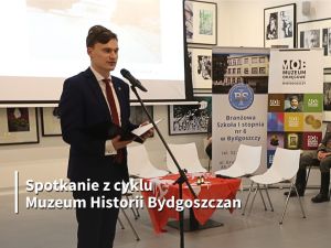 Młody mężczyzna przed mikrofonem. W tle duży ekran i obrazy.