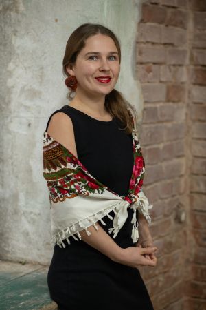 Uśmiechnięta długowłosa młoda kobieta w czarnej sukience z kolorową chustą ludową na ramionach.