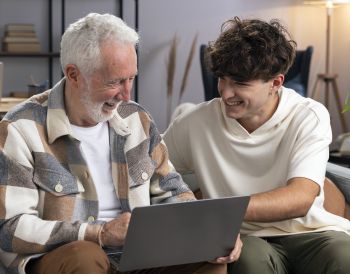 Starszy mężczyzna z laptopem na kolanach i młody chłopak siedzą roześmiani na kanapie.
