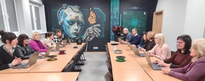 Osoby przy stolikach z laptopami. W tle ściana z muralem przedstawiającym humanoida.