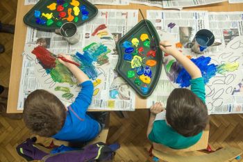 Zdjęcie robione z góry, dwaj chłopcy malują pędzlami, obok palety z kolorowymi farbami.