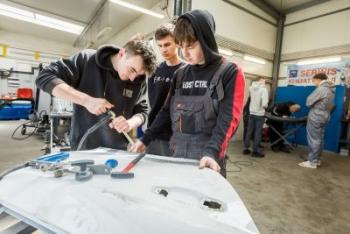 Uczniowie z narzędziami w warsztacie samochodowym pochyleni nad karoserią samochodu.