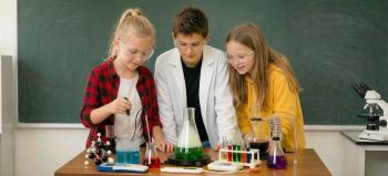 Troje uczniów w goglach ochronnych przeprowadza eksperyment chemiczny w klasie.