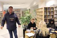 Mężczyzna w niebieskiej kurtce pozuje do zdjęcia przy osobach siedzących przy stole