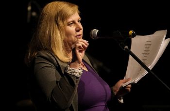 Kobieta o jasnych długich włosach przy mikrofonie na scenie, w ręku trzyma kartki.