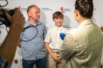 Uśmiechnięty chłopiec udziela wywiadu na tle baneru Woj. Kujawsko-Pomorskiego, obok stoi mężczyzna.