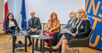 Uczestnicy debaty siedzą w fotelach przy stolikach. Kobieta trzyma mikrofon.