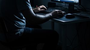 Ręce mężczyzny na klawiaturze komputerowej w ciemnym pokoju.