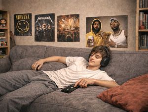 Nastolatek z zamkniętymi oczami i słuchawkami leży na kanapie ze smartfonem. W tle plakaty muzyków.