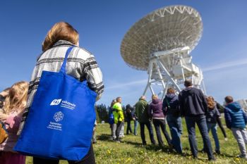 Ludzie stoją pod wielkim radio teleskopem na łące.