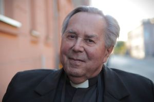 Siwowłosy, starszy mężczyzna w ciemnym ubraniu z koloradką.