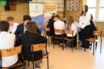 Grupa młodzieży w białych koszulach siedzi przy dwóch stolikach, przy jednym z nich stoi uczennica i wypowiada się.