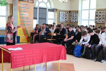 Dorośli i młodzież siedzący na krzesłach patrzą na kobietę przemawiającą do nich przy stole.