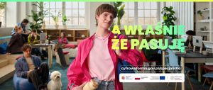 Młoda kobieta na pierwszym planie w nowoczesnym pomieszczeniu, w tle pracujący przy komputerach inne kobiety i mężczyźni.