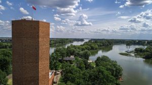 Wieża z cegieł z flagą biało-czerwoną na szczycie i jezioro w tle.